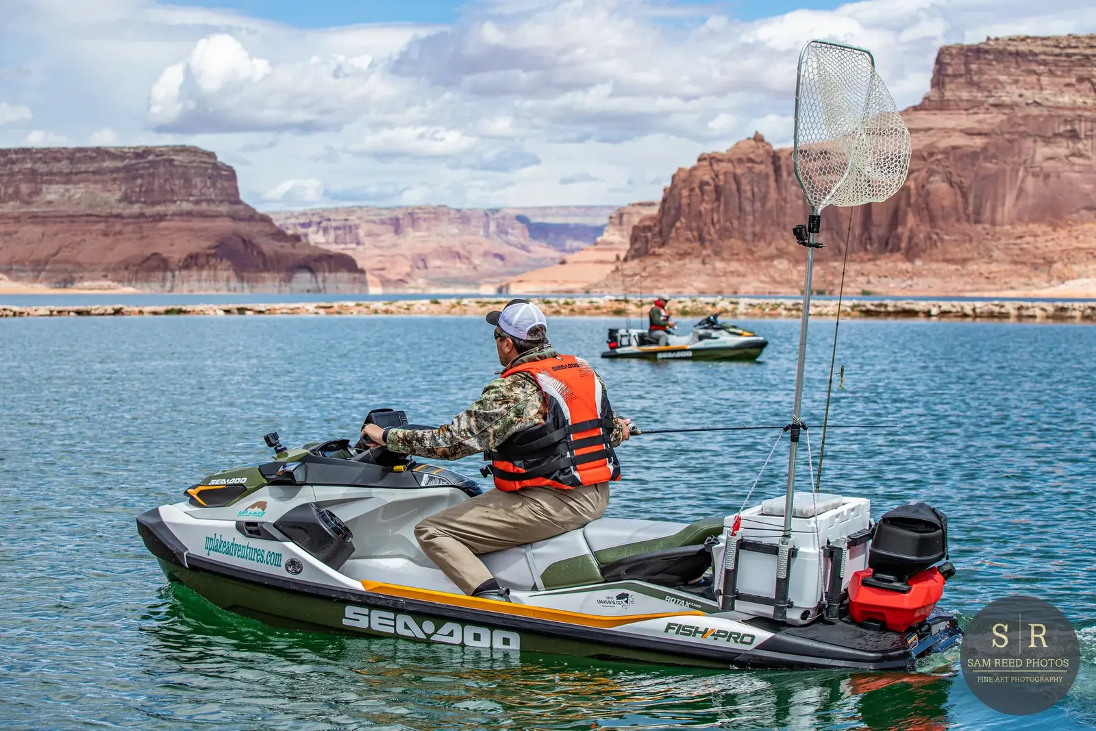 Person on a Sea-Doo watercraft fishing with a net attached to a pole on a lake with red rock formations in the background.