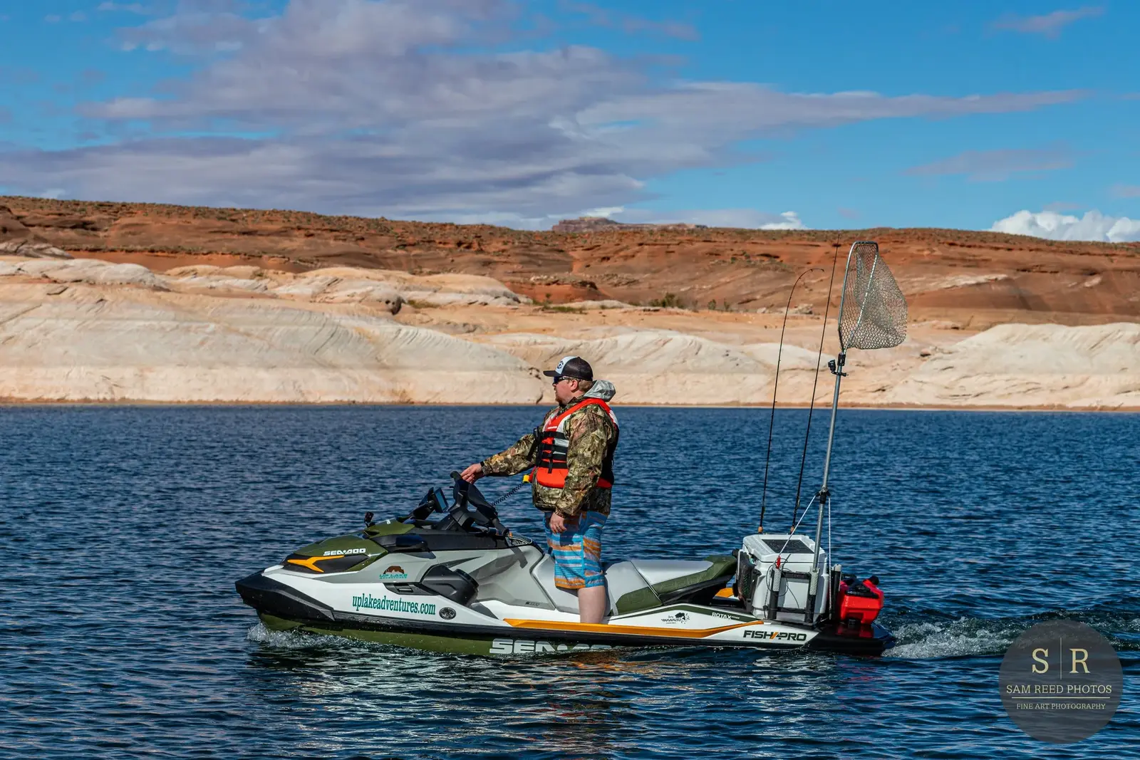 A man standing on a jet ski on a lake, with a fishing net and gear attached, surrounded by canyon walls and a partly cloudy sky.