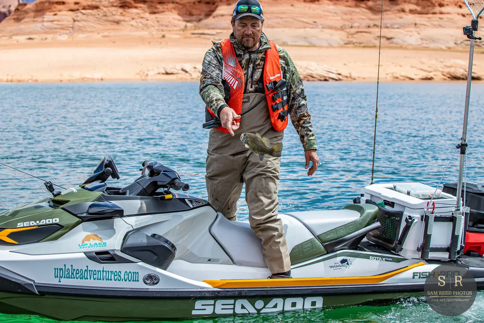 A man in camouflage clothing and an orange life vest is standing on a motorized fishing kayak on a lake, holding a small fish he caught.