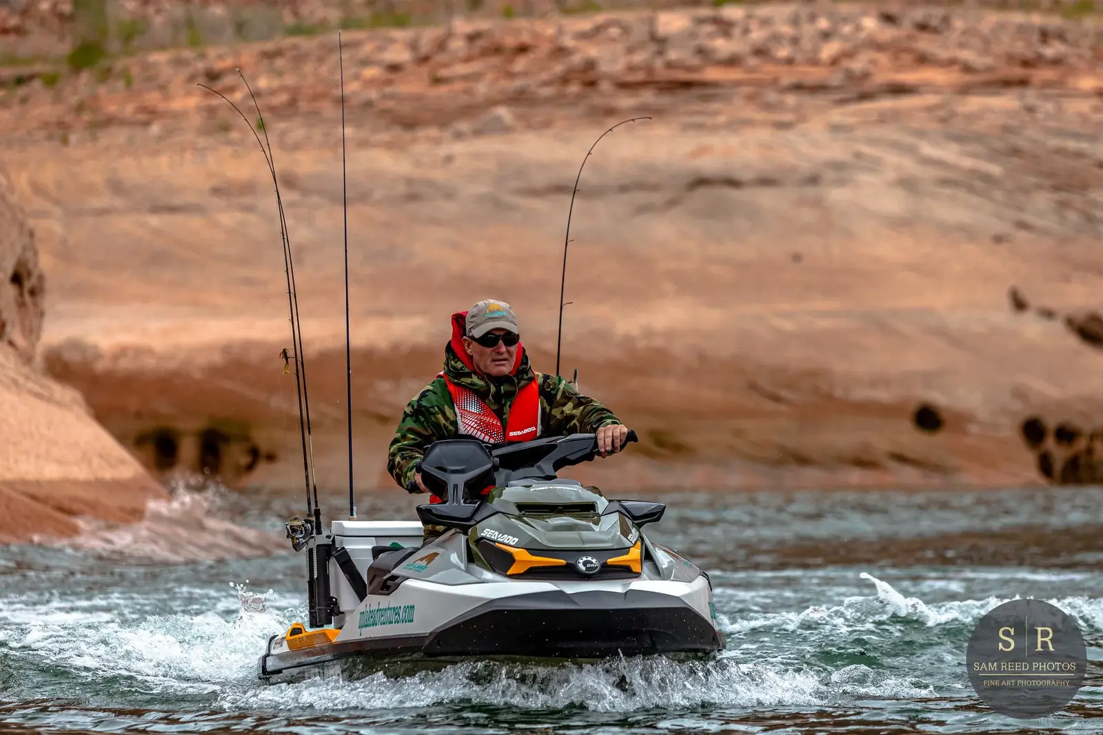 Man in camouflage jacket and sunglasses riding a jet ski on water with rocky canyon background.