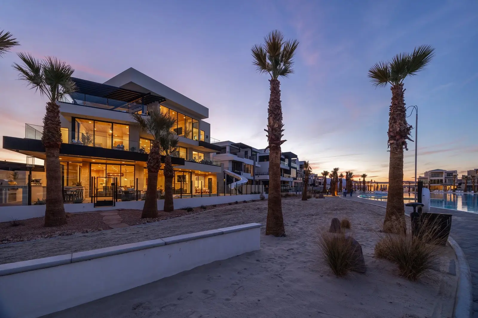 Angled view of a glowing modern home and palm-lined path at twilight