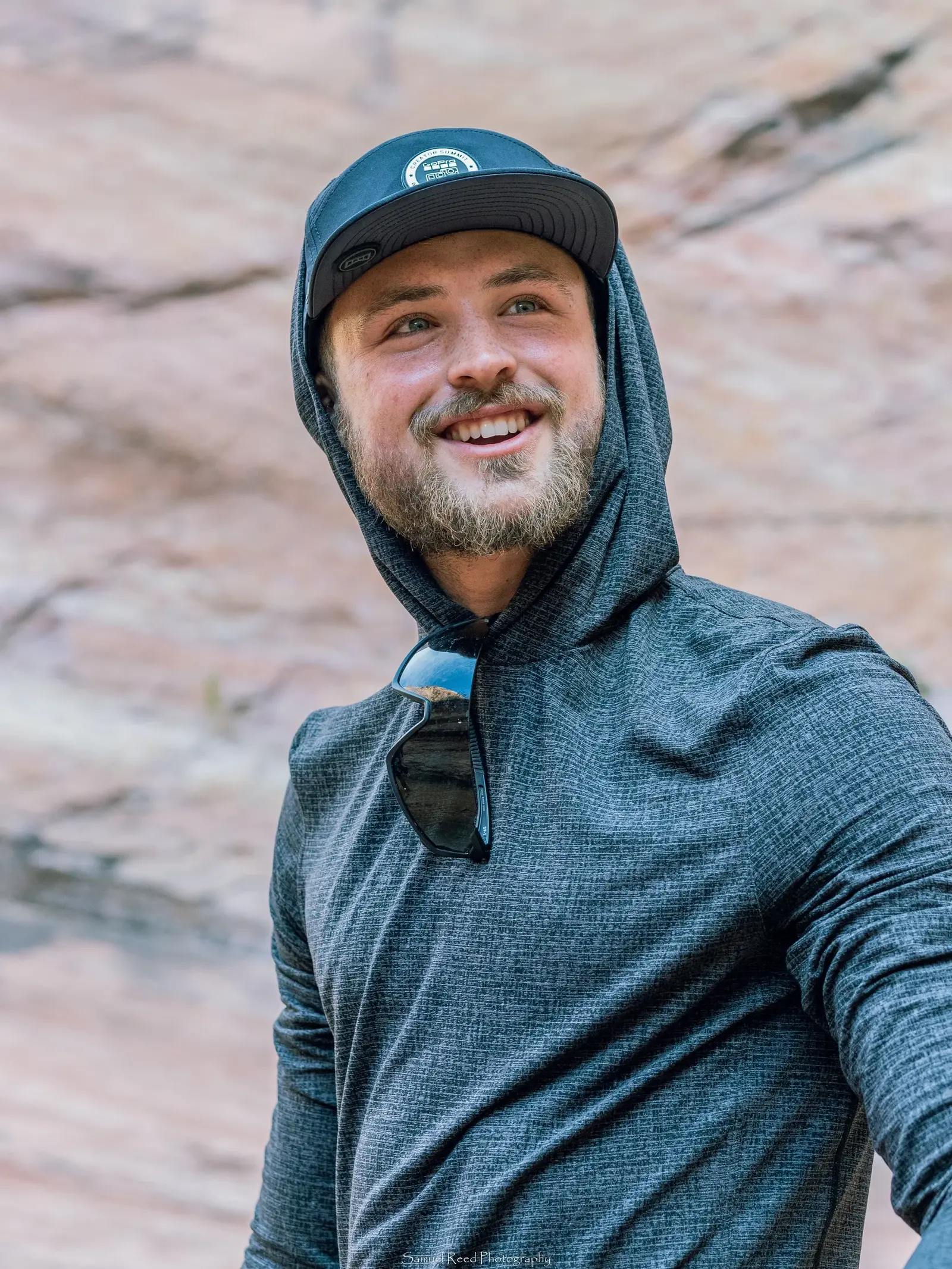 A smiling man in a gray hoodie and black cap outdoors with a rocky background.