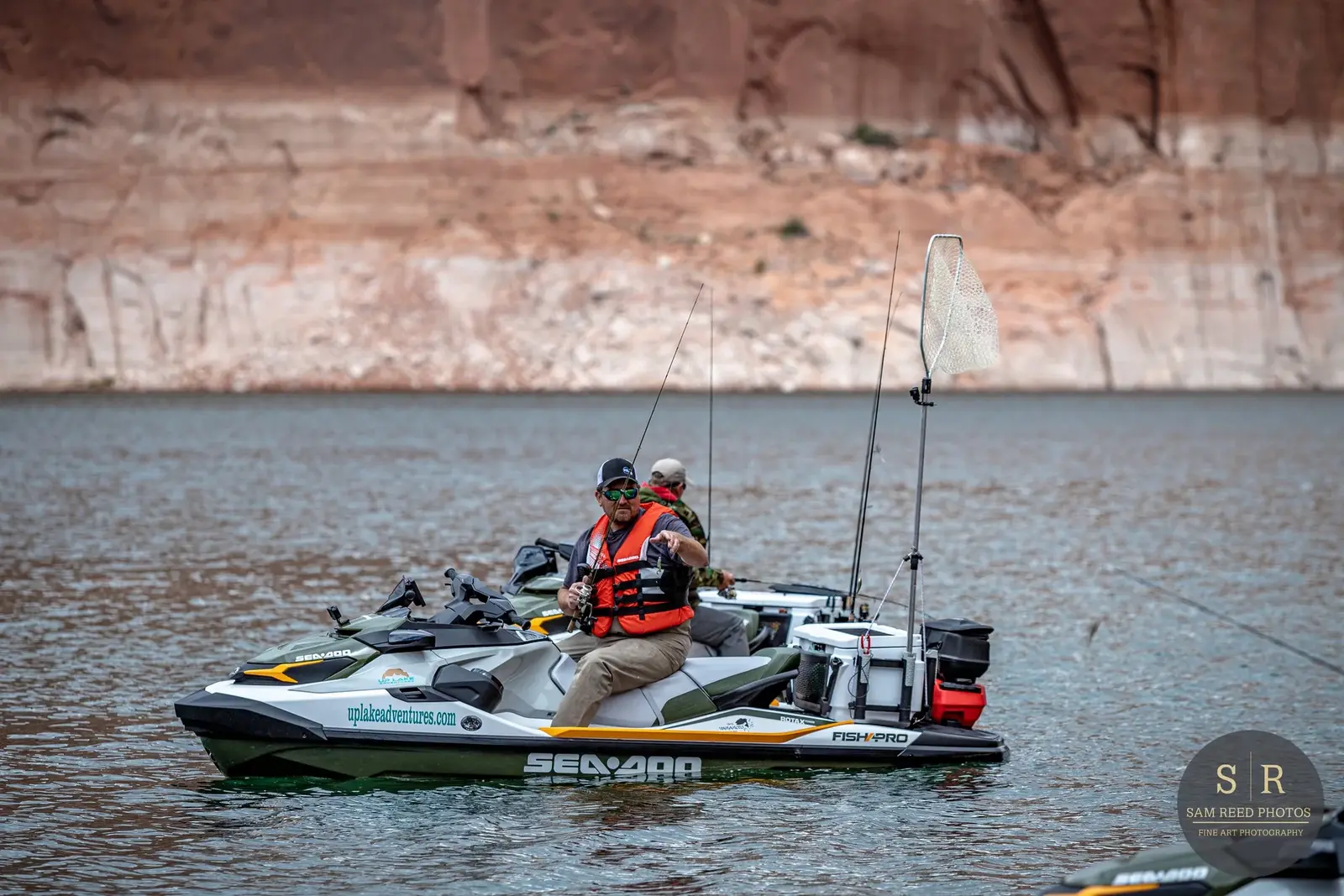 Two men fishing on a jet ski on a lake with rocky cliffs in the background.
