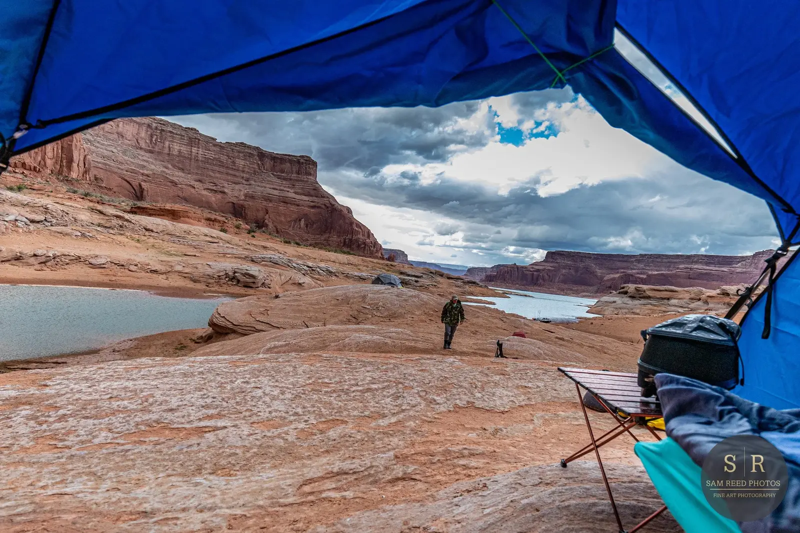 View from inside a blue camping tent overlooking a river and red rock formations in a canyon, with a person walking along the rocky terrain and cloudy sky.