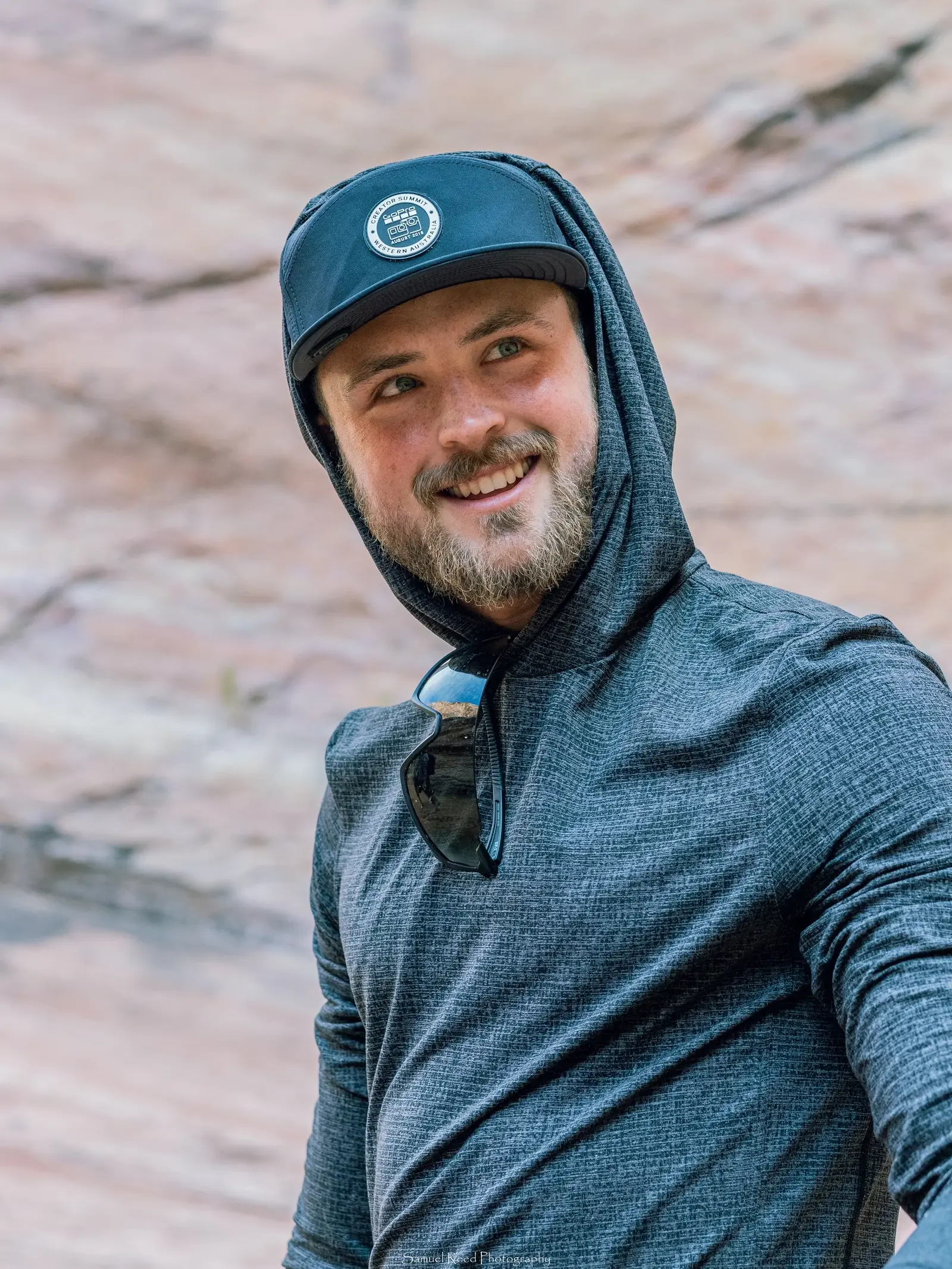 A man with a beard smiling, wearing a black hooded jacket, a blue cap with a circular logo, and sunglasses hanging from his collar, with a rocky background.