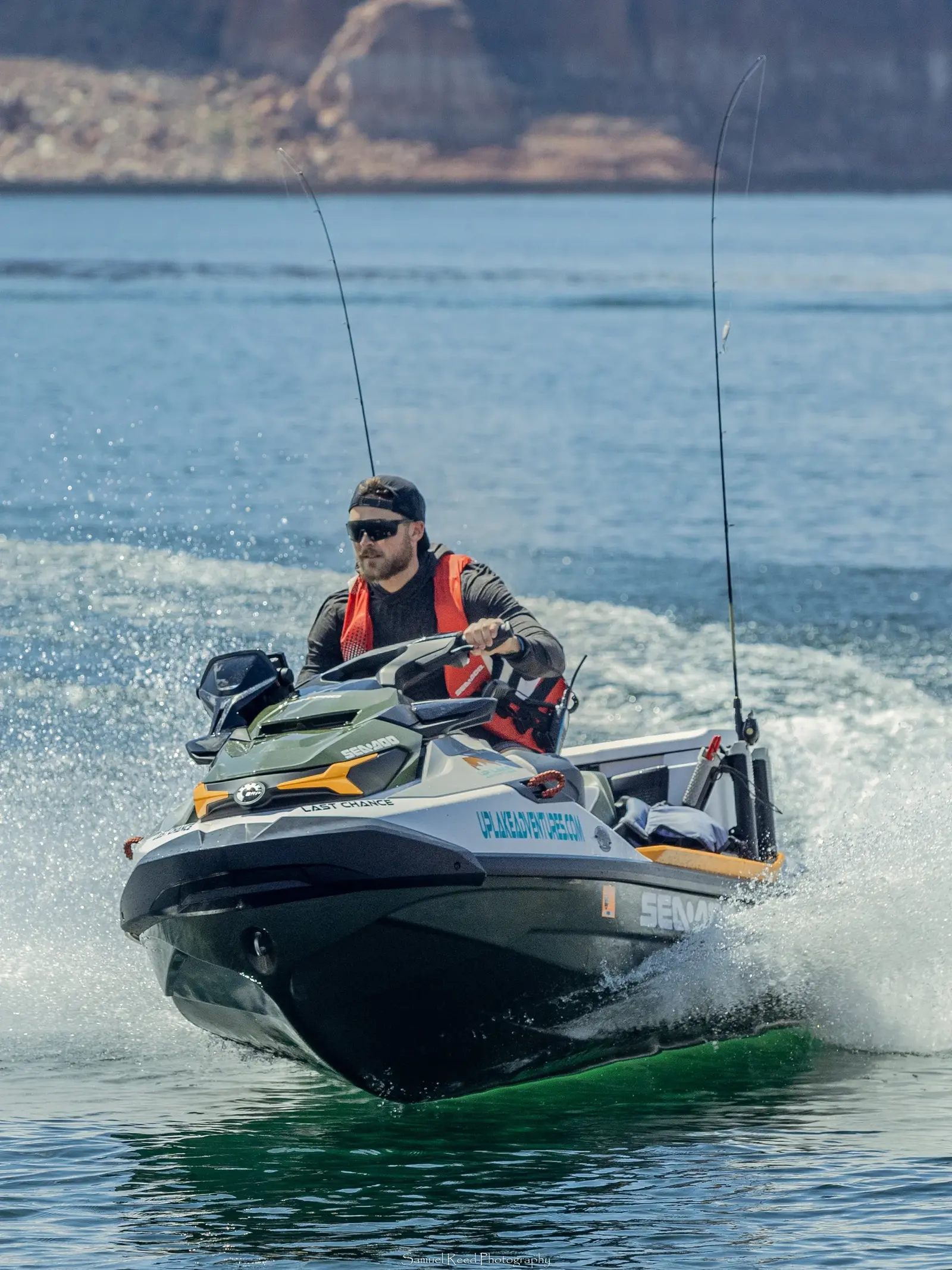 A man wearing sunglasses, a cap, and an orange life vest rides a jet ski on a body of water with rocky cliffs in the background.
