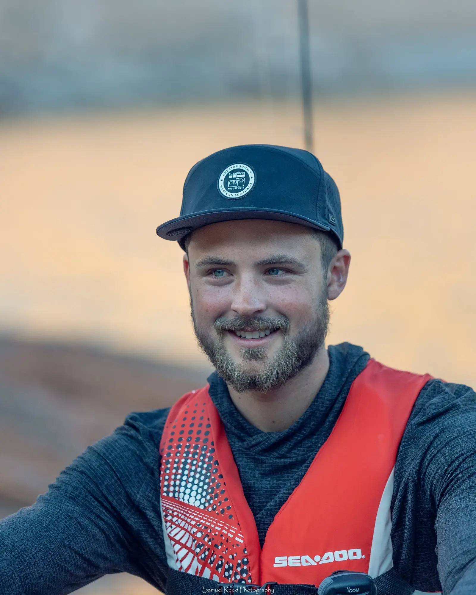 A smiling man wearing a baseball cap and an orange life vest outdoors during sunset.