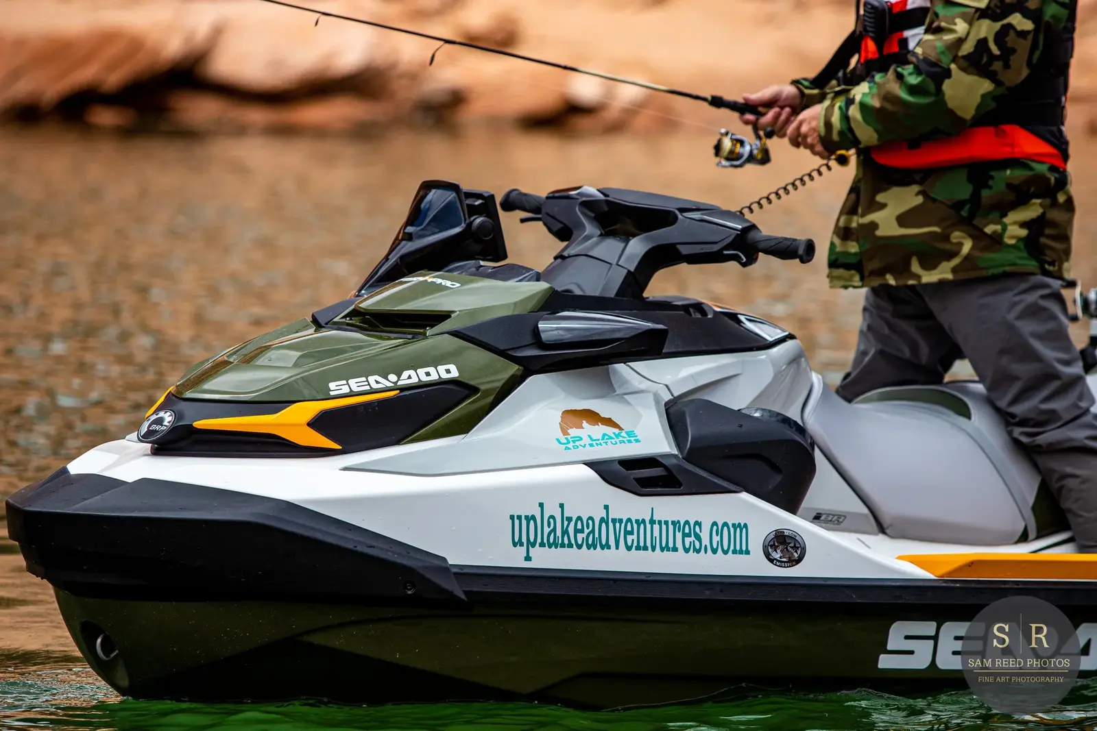 A person in camouflage clothing riding a green and white Sea-Doo jet ski on a lake with a rocky shoreline in the background.