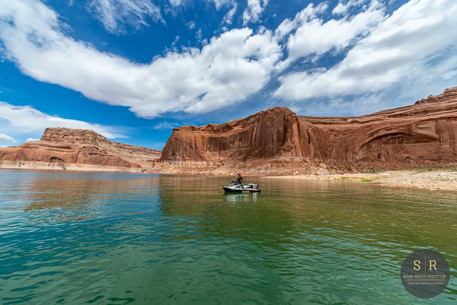 Person on a jet ski on a lake with red rock cliffs and blue sky with white clouds in the background.