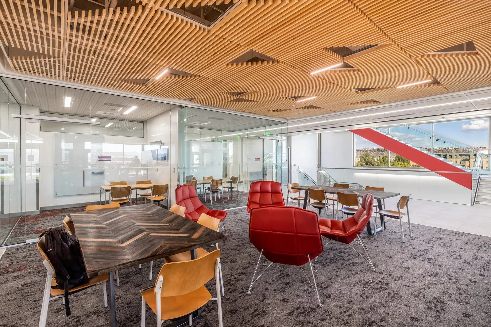 Shared study area beneath a geometric timber ceiling, with red lounge chairs and a glass-walled stair.