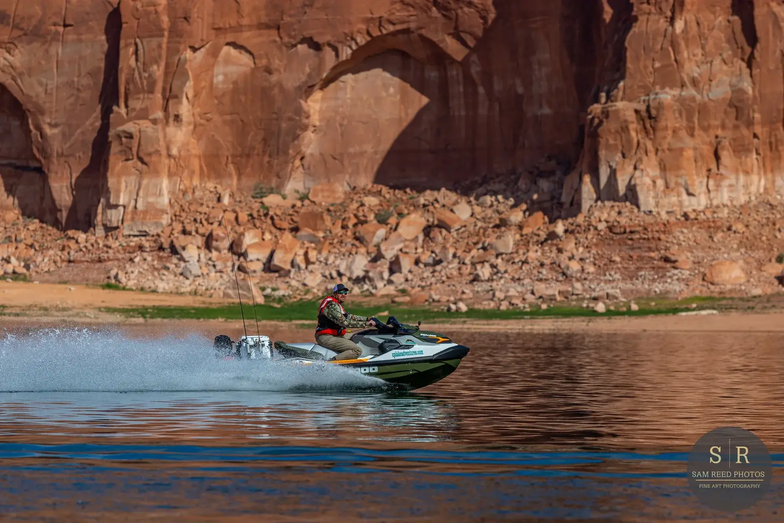 A man riding a jet ski on a body of water with a rocky red canyon and large arch in the background.