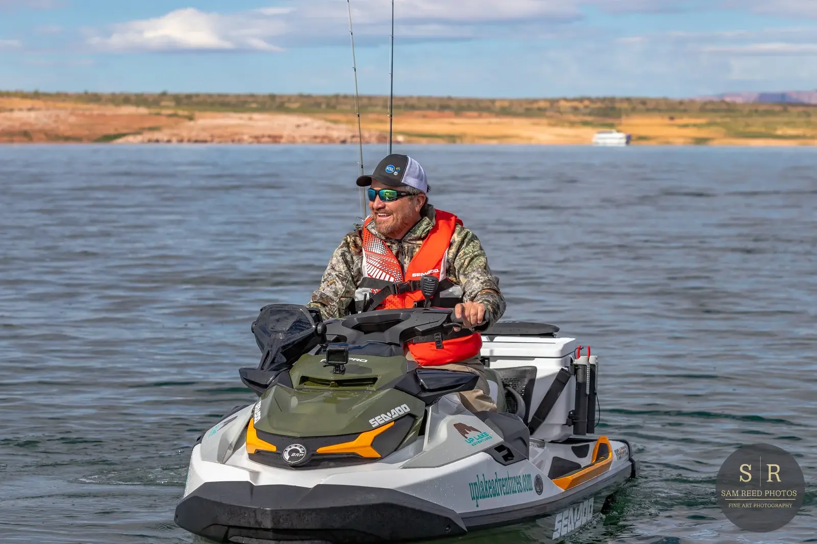 Man in camouflage jacket and red life vest riding a yellow and white Sea-Doo jet ski on a lake with distant shoreline and boat in the background.