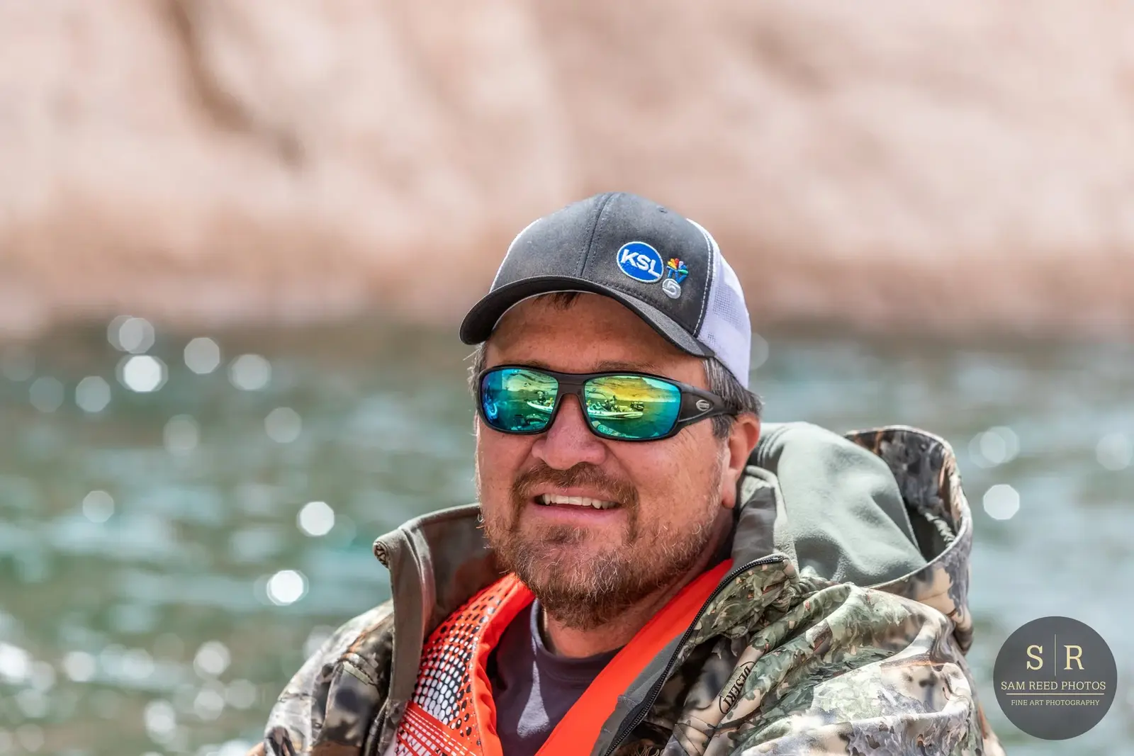 A man wearing sunglasses, a baseball cap with a 'KSL TV' logo, a camouflage jacket, and an orange life vest, standing near water with a rocky background.