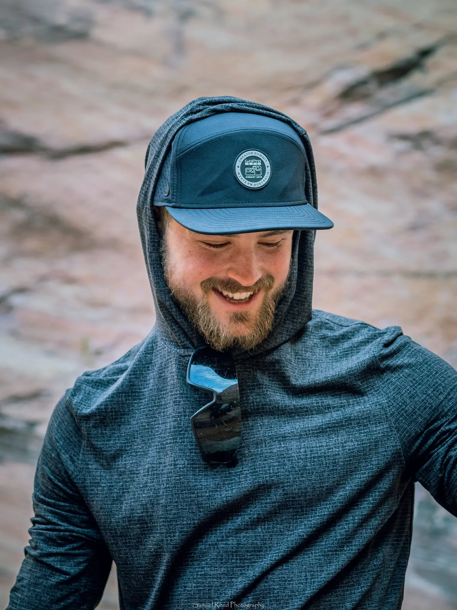 A man smiling outdoors wearing a blue cap, a gray hoodie with the hood up, and sunglasses hanging from his shirt.