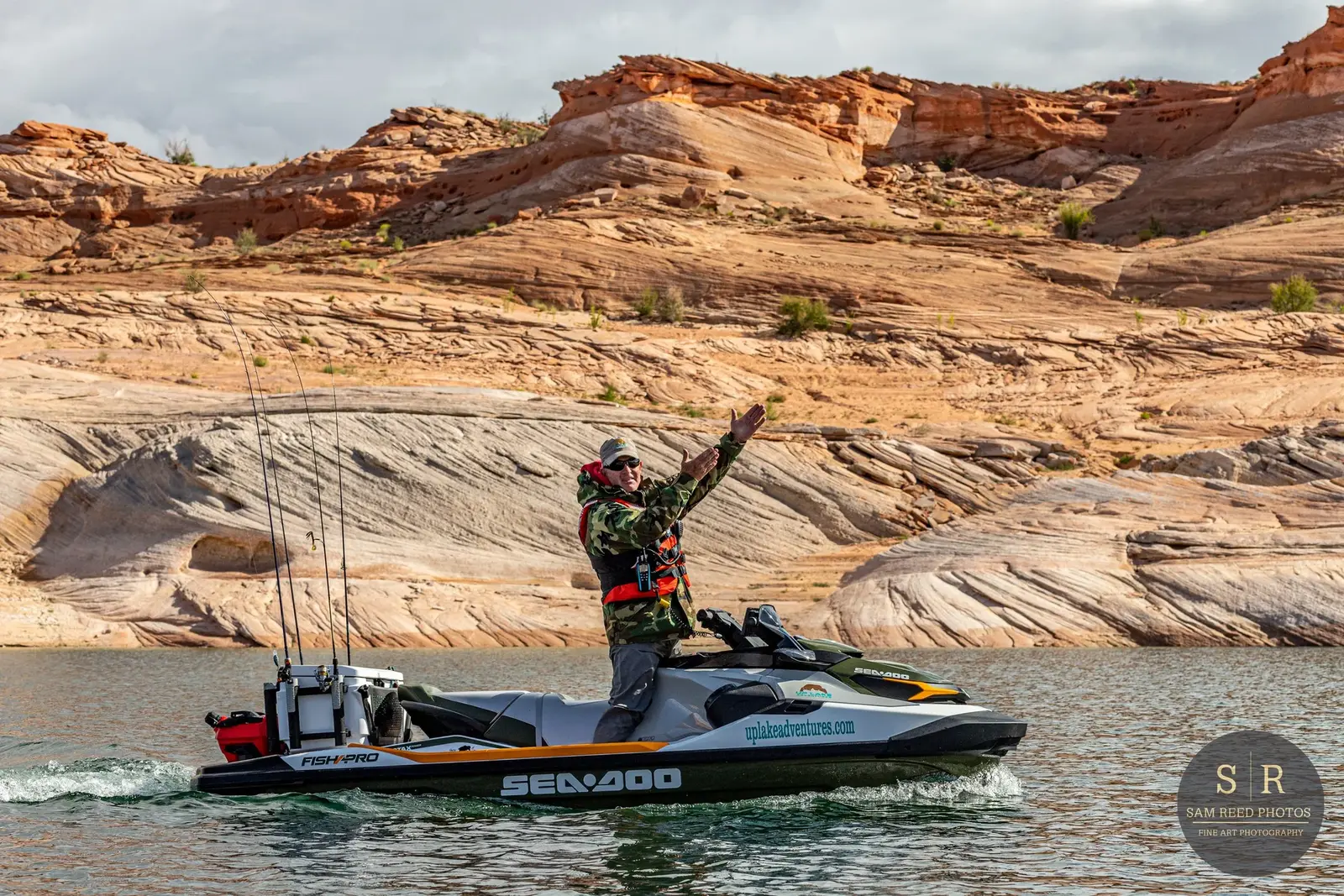 A man in camouflage jacket and sunglasses stands on a Sea-Doo jet ski on a lake with rocky red sandstone cliffs in the background.