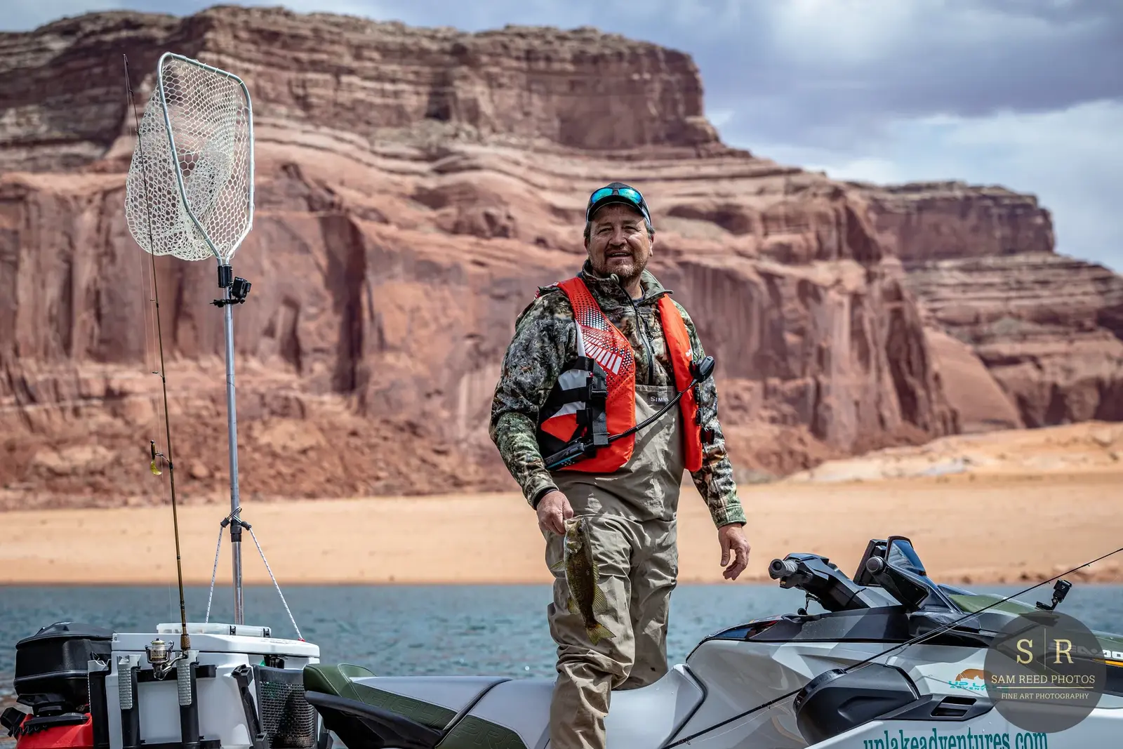 A man standing on a jet ski holding a fish, with a lake and redrock cliffs in the background.