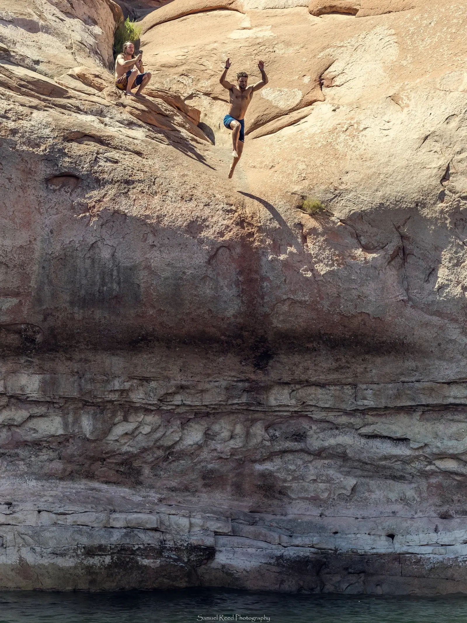 A shirtless man jumping off a cliff into water below, with a person sitting nearby taking a photo against a rocky cliff background.
