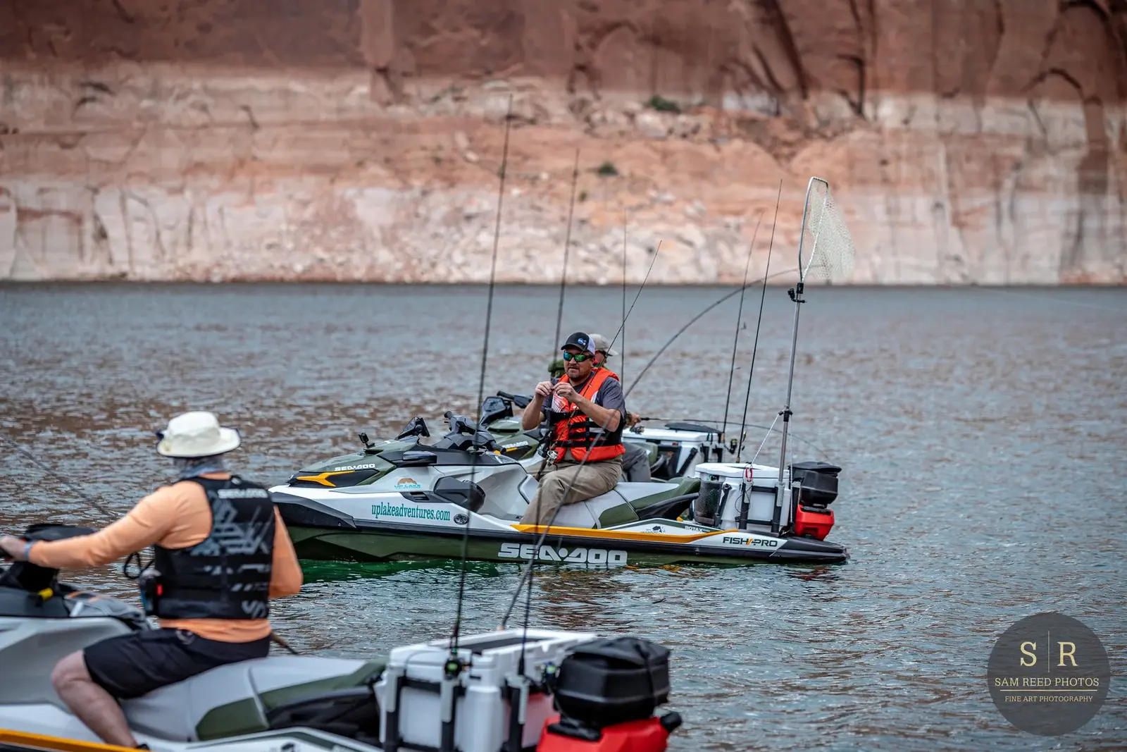 Two people on jet skis on a lake, one man fishing with a rod, in front of a rocky canyon background.