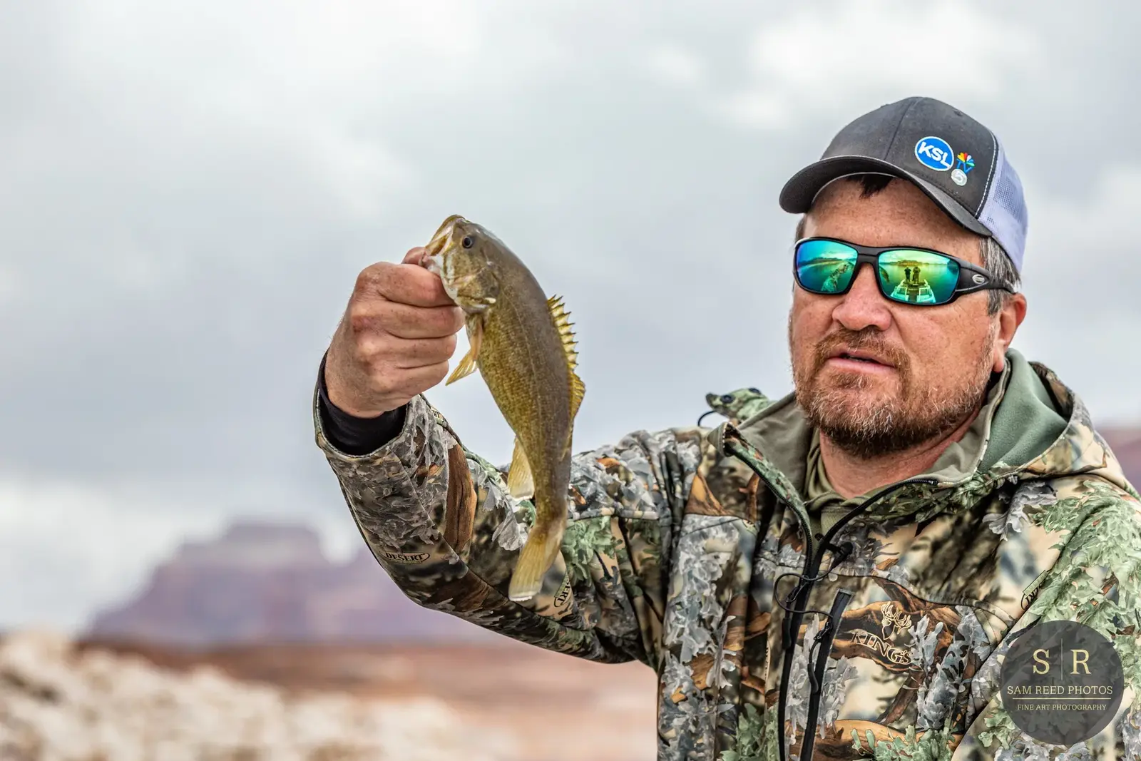 A man wearing camouflage jacket, sunglasses, and a cap, holding a small fish outdoors with a landscape of mesas and cloudy sky in the background.