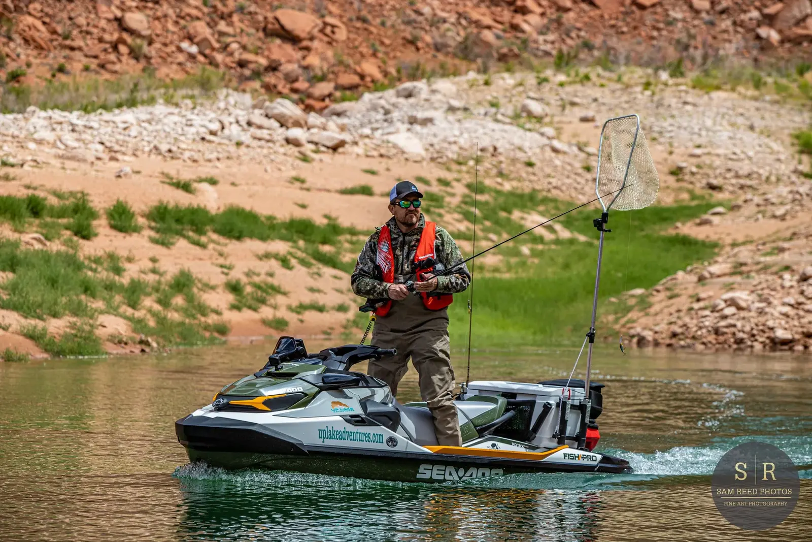 Man fishing on a jet ski in a river with green and rocky landscape in the background.