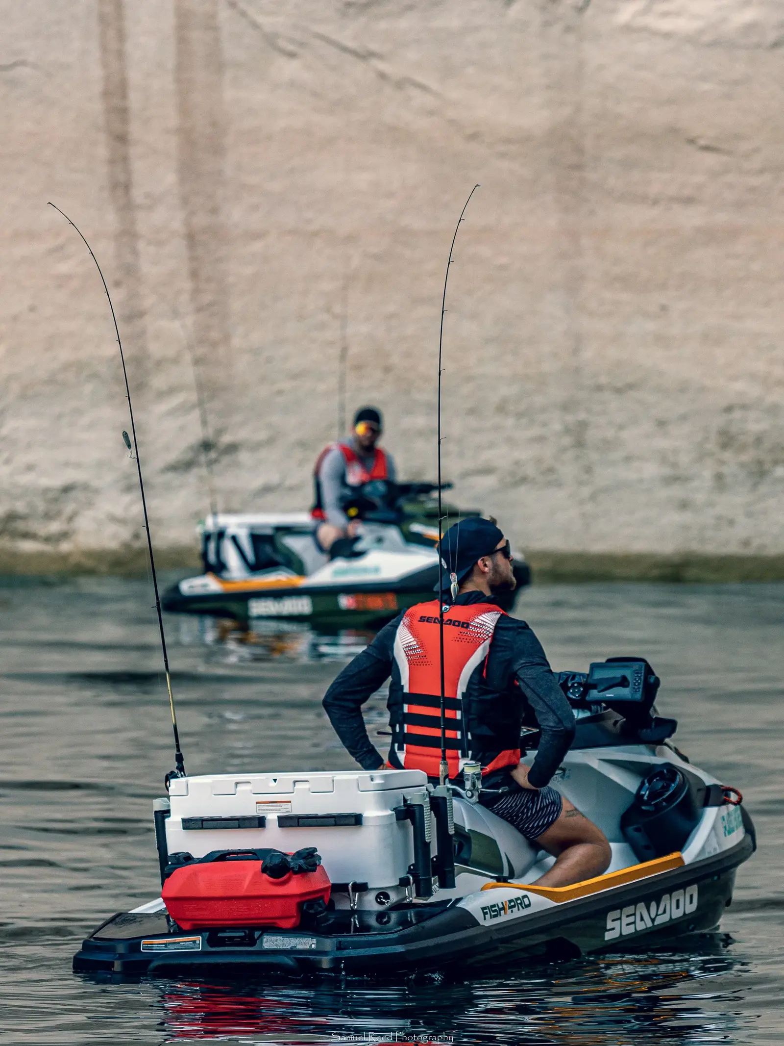 Two men riding on small motorized boats on a body of water, with a sandy hill in the background. The men are wearing life jackets and sunglasses.