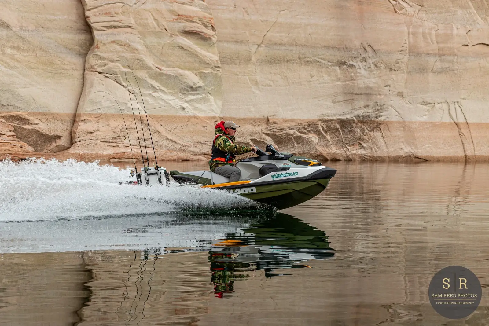 A man wearing camouflage jacket and red hoodie driving a white and green jet boat on calm water near rocky cliffs, with three fishing rods mounted on the boat.
