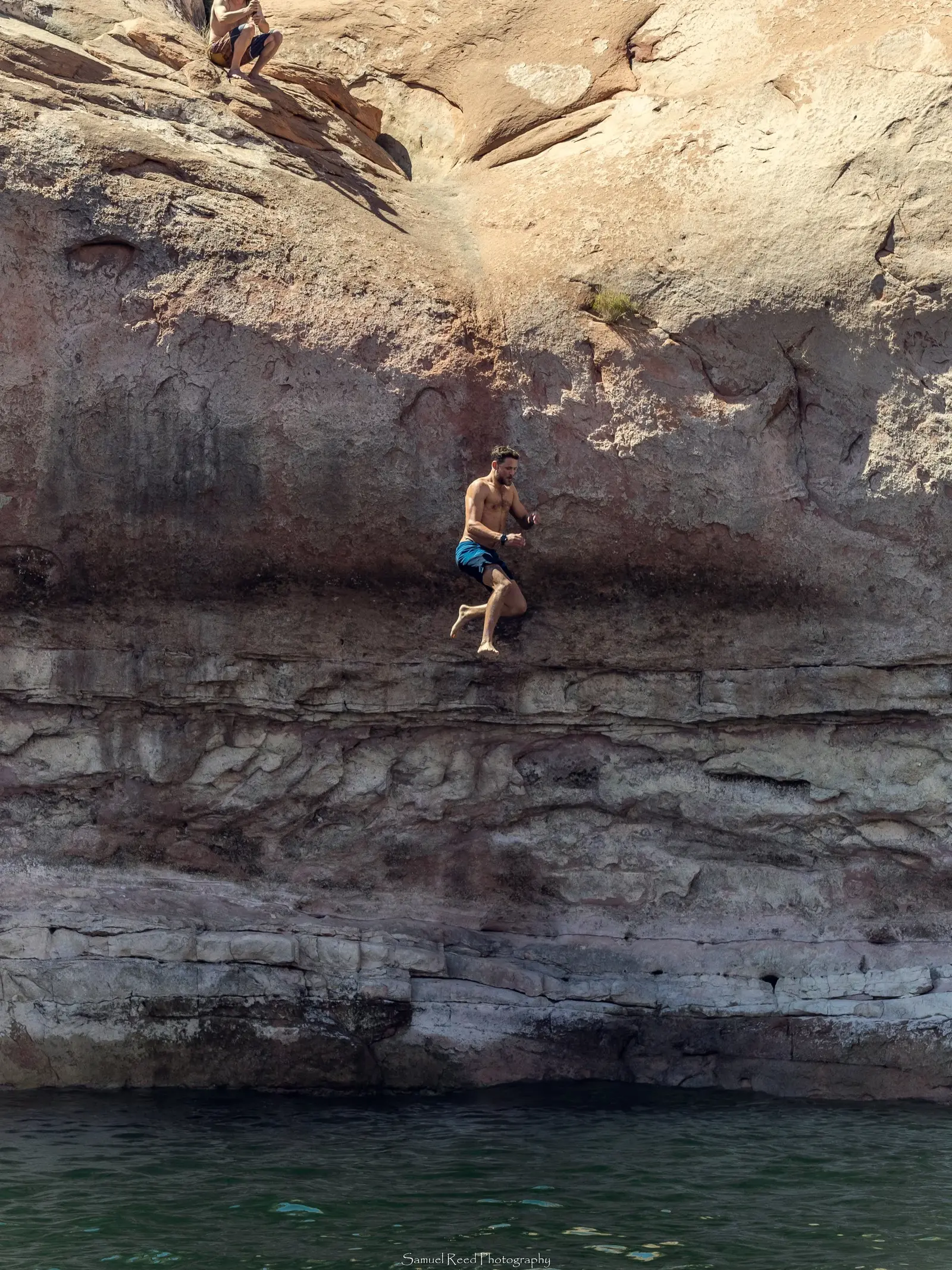 A man in blue swim trunks jumping off a rocky ledge into the water below, with another person sitting on the rocks above.