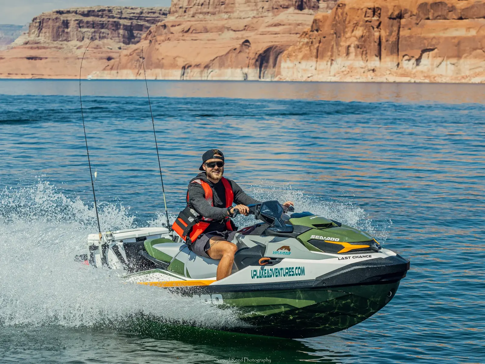 Man riding a green and black SEA-DOO jet ski on a lake with canyon walls in the background, wearing sunglasses, a black cap backward, a red life jacket, and a long-sleeved dark shirt.