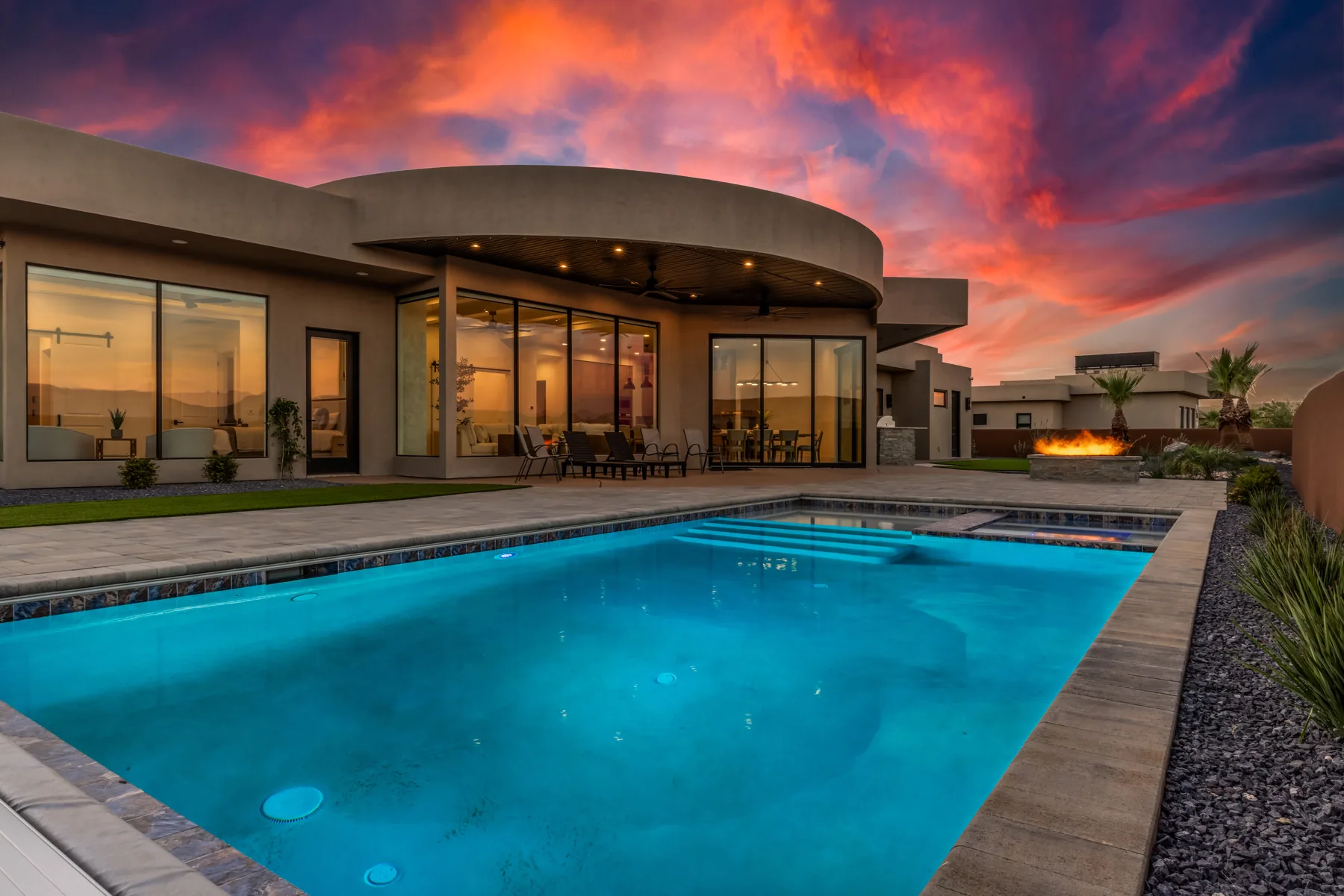 Illuminated pool and curved-roof home beneath an orange and purple twilight sky